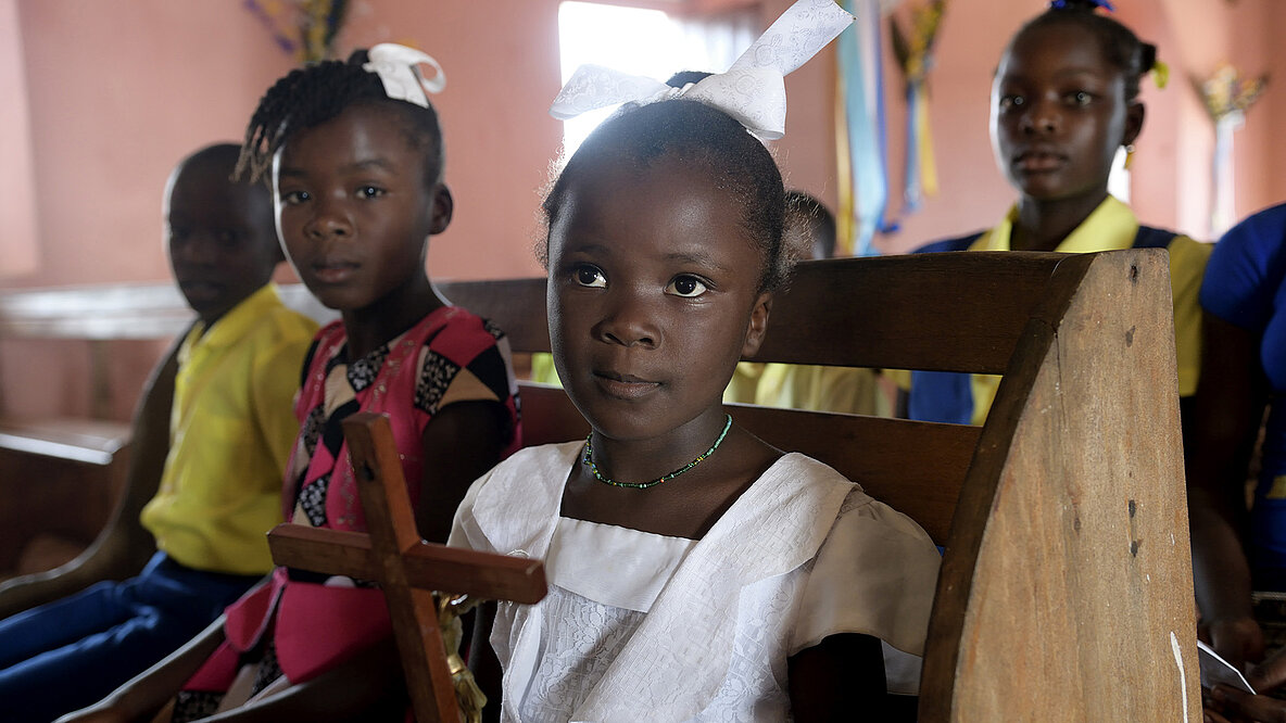 © Florian Kopp / Kindermissionswerk Ein Mädchen aus Haiti sitzt mit einem Kreuz in der Kirchenbank